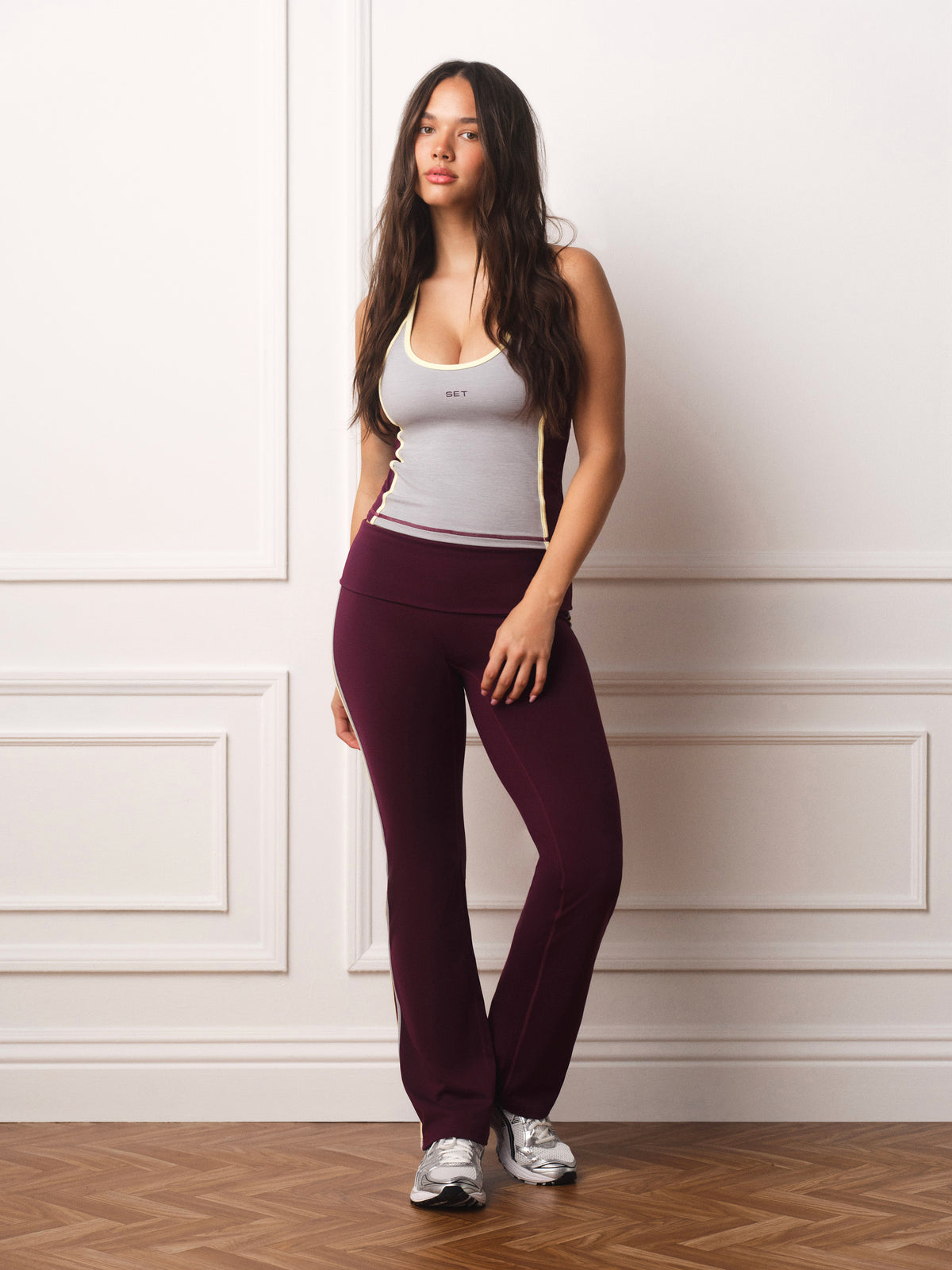 A woman stands against a white paneled wall wearing dark burgundy athletic pants, a light gray tank top, and silver sneakers. <model-info>Leah wears a size M and is 5’7”, 25” waist, 32D bust.</model-info>