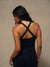A woman shown from the back wearing a black athletic tank top with crisscross shoulder straps and matching black leggings, standing against a neutral textured beige wall.