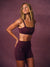 A woman modeling a matching burgundy activewear set consisting of a sports bra and high-waisted shorts, posed against a textured mauve studio backdrop.