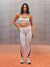 A woman modeling a matching white activewear set with black trim, featuring a sports bra and high-waisted leggings, standing against a textured gray background with a terracotta-colored floor. <model-info>Alana wears a size M and is 5’8”, 28” waist, 35" bust.</model-info>
