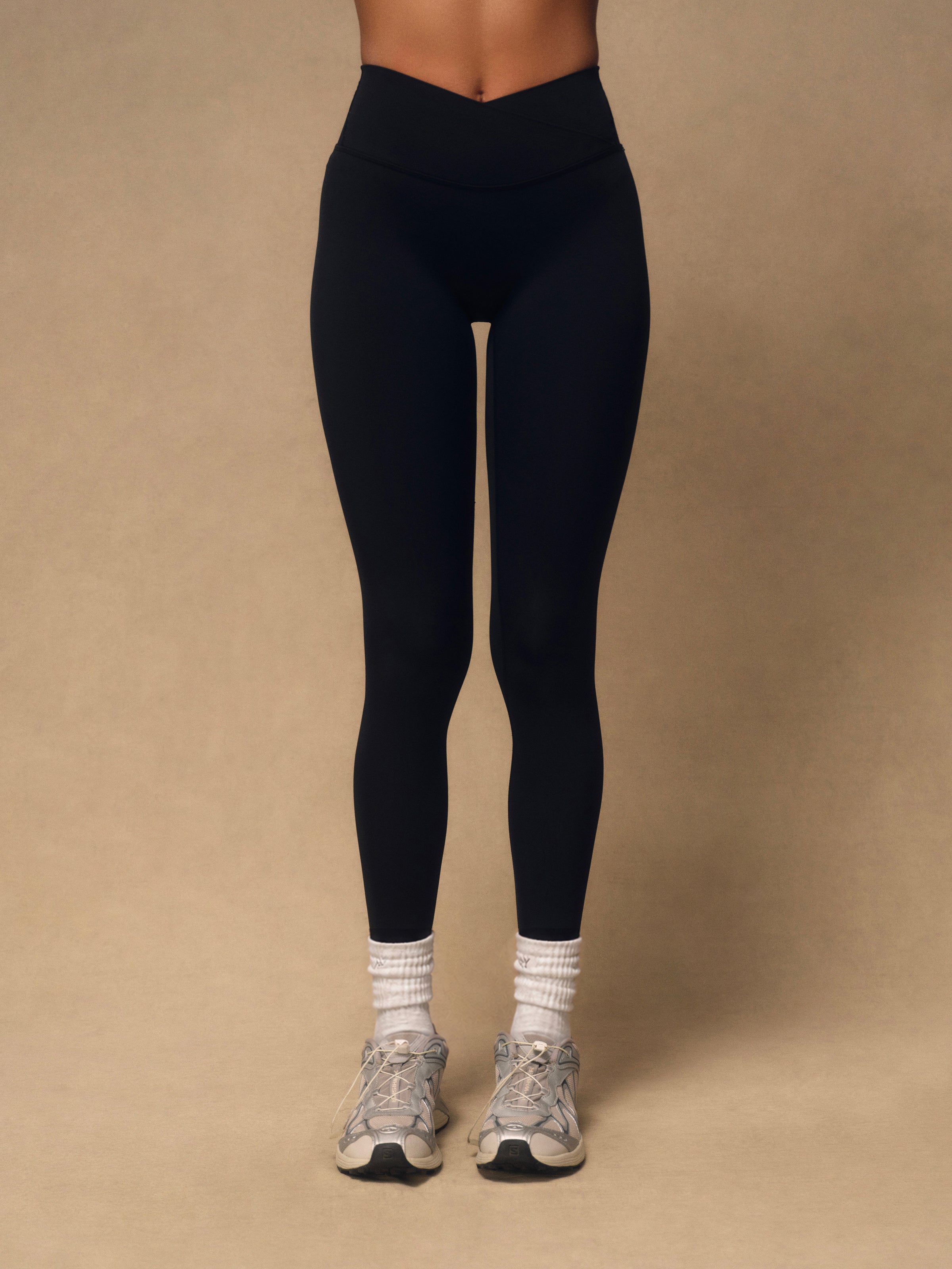 Black high-waisted leggings worn by a model, paired with white socks and gray sneakers, standing against a solid beige studio background.