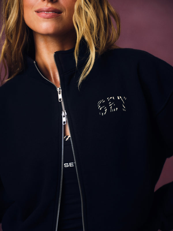 Close-up of a woman wearing a dark navy zip-up jacket with a white textured 'SET' logo on the chest, set against a solid mauve background.