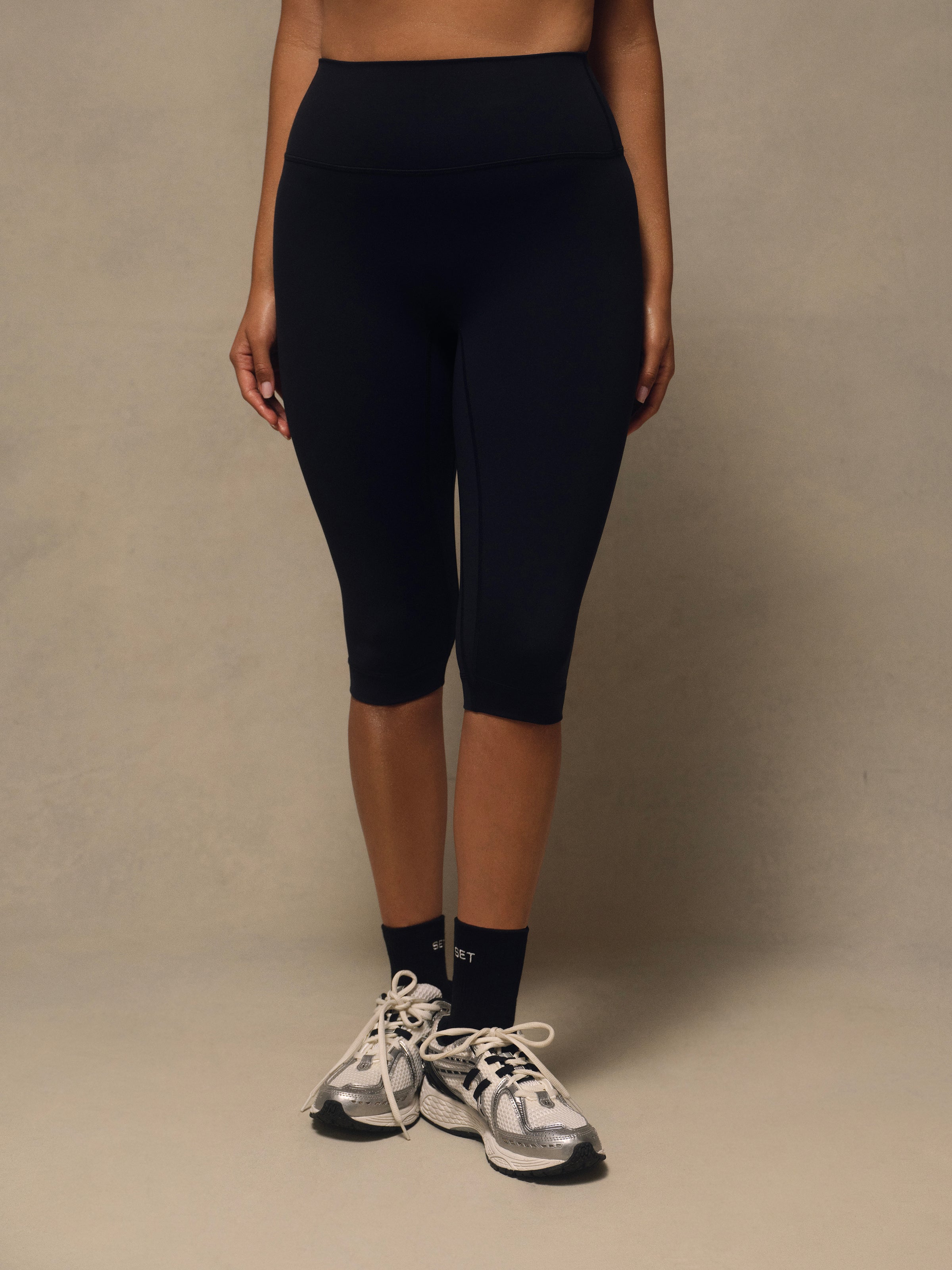 A model wearing black high-waisted capri leggings, styled with black crew socks and gray and white running shoes, standing against a neutral beige studio backdrop.
