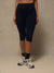 A model wearing black high-waisted capri leggings, styled with black crew socks and gray and white running shoes, standing against a neutral beige studio backdrop.