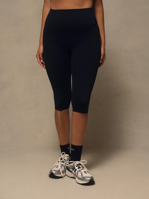 A model wearing black high-waisted capri leggings, styled with black crew socks and gray and white running shoes, standing against a neutral beige studio backdrop.
