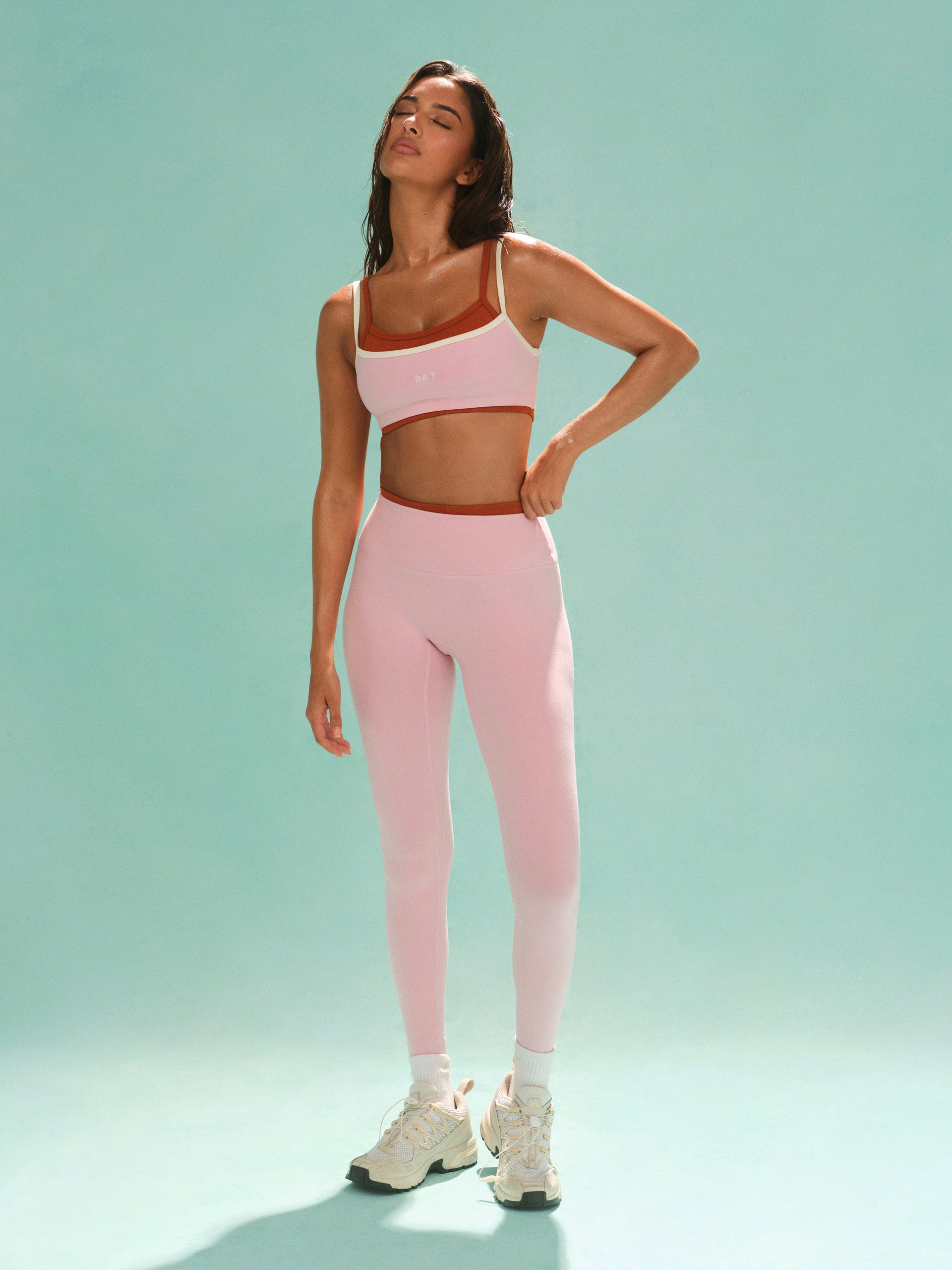 Woman wearing a pink athletic outfit on a light green background <model-info>Kushi wears a size S and is 5'8, 24” waist, 32C bust.</model-info>