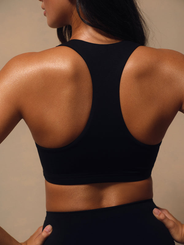 Rear view of a woman wearing a black racerback sports bra and matching black bottoms against a warm beige studio background.