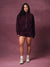 A woman modeling a matching burgundy zip-up hoodie and shorts set against a textured mauve studio backdrop.