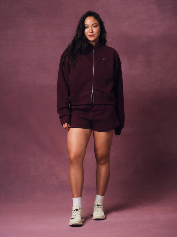 A woman modeling a matching burgundy zip-up hoodie and shorts set against a textured mauve studio backdrop.
