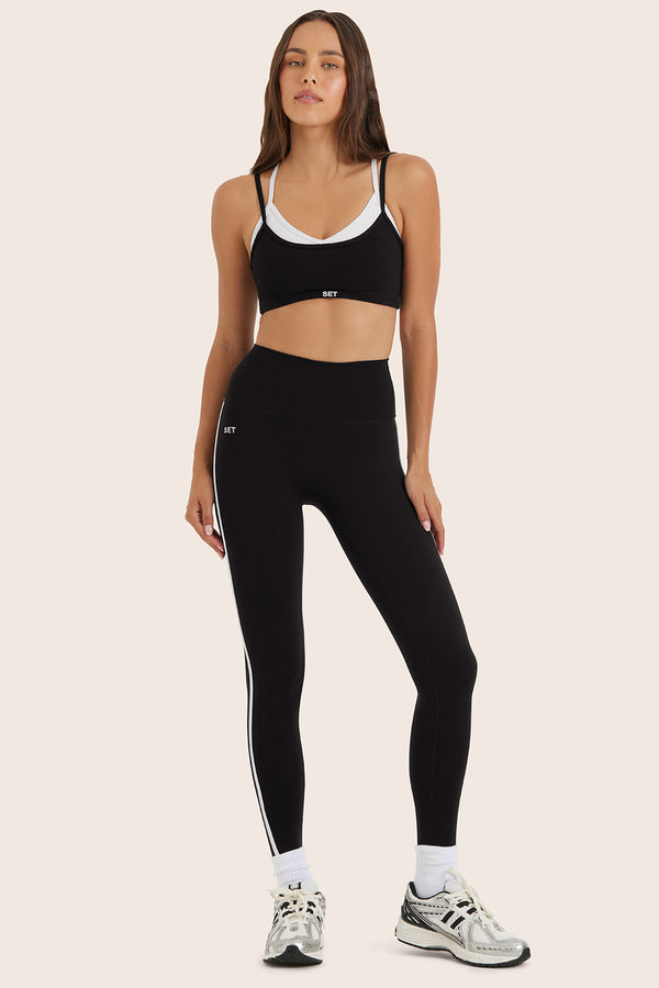 Woman wearing black sports bra and leggings with white accents on a beige background