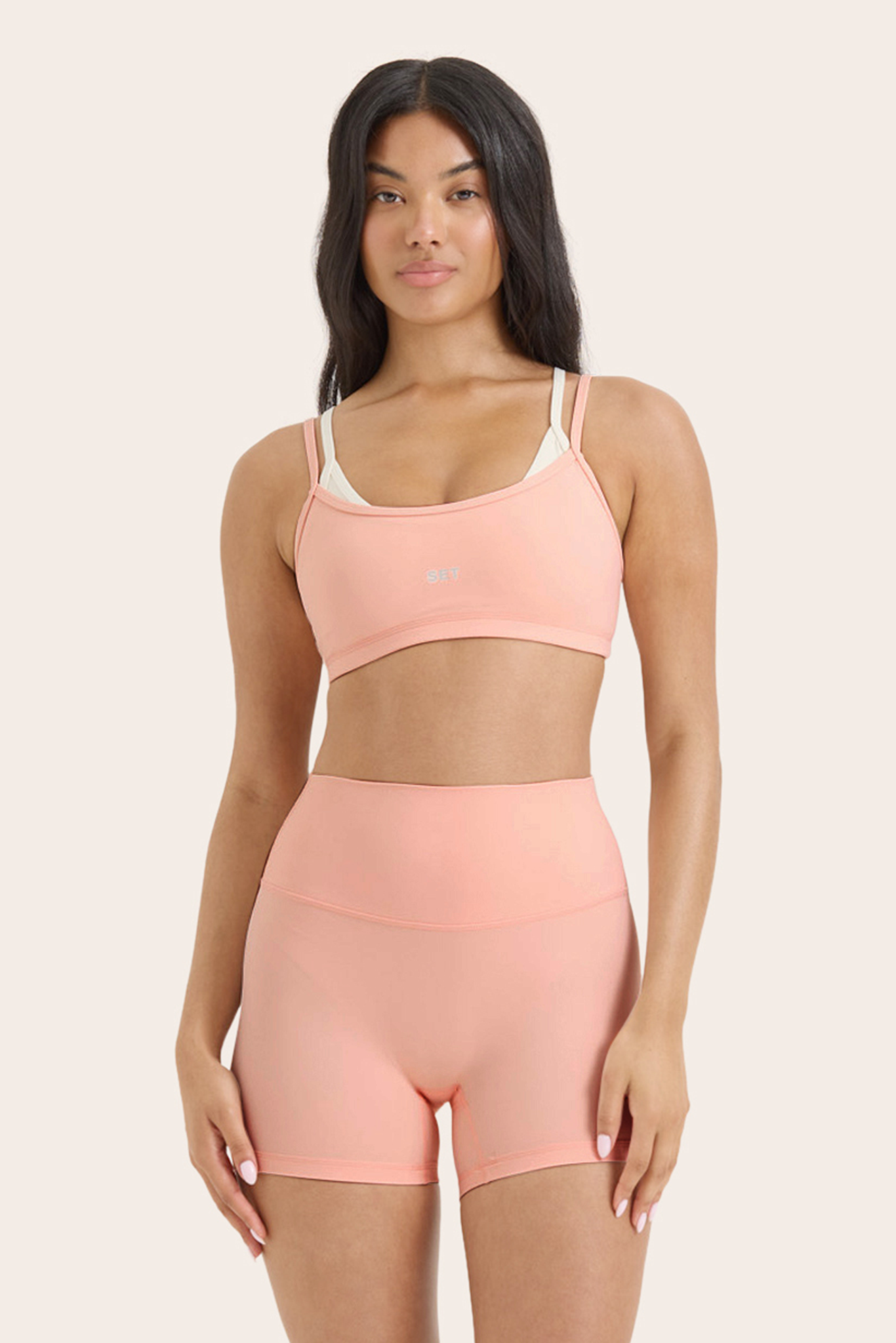 Woman wearing a peach-colored sports bra and shorts set on a beige background 