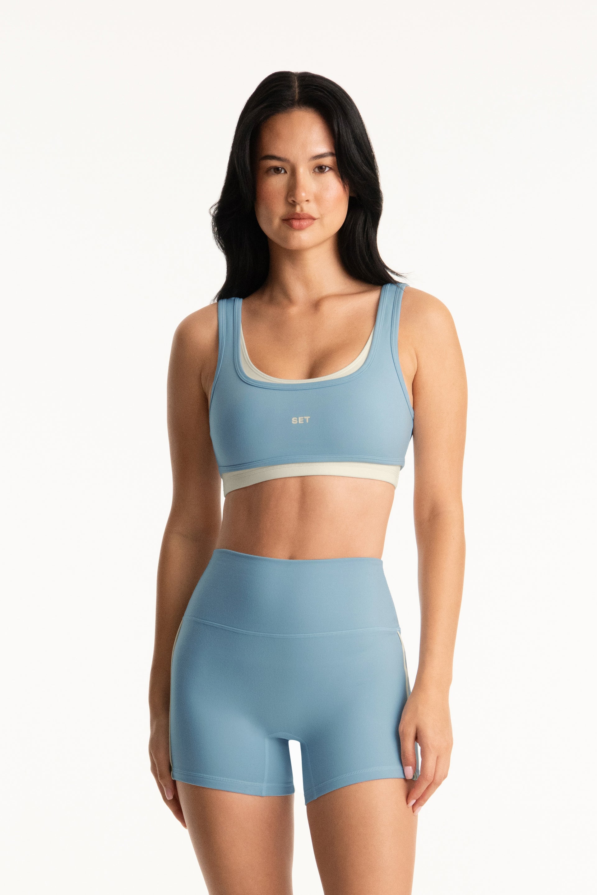 Woman wearing a blue sports bra and shorts set on a white background <model-info>Jasmine wears a size S and is 5’8”, 25" waist, 36” bust.</model-info>
