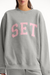 Gray sweatshirt with pink 'SET' letters worn by a person on a plain background