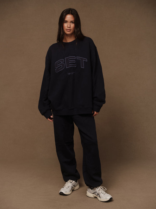 Woman wears navy sweat suit in front of brown background