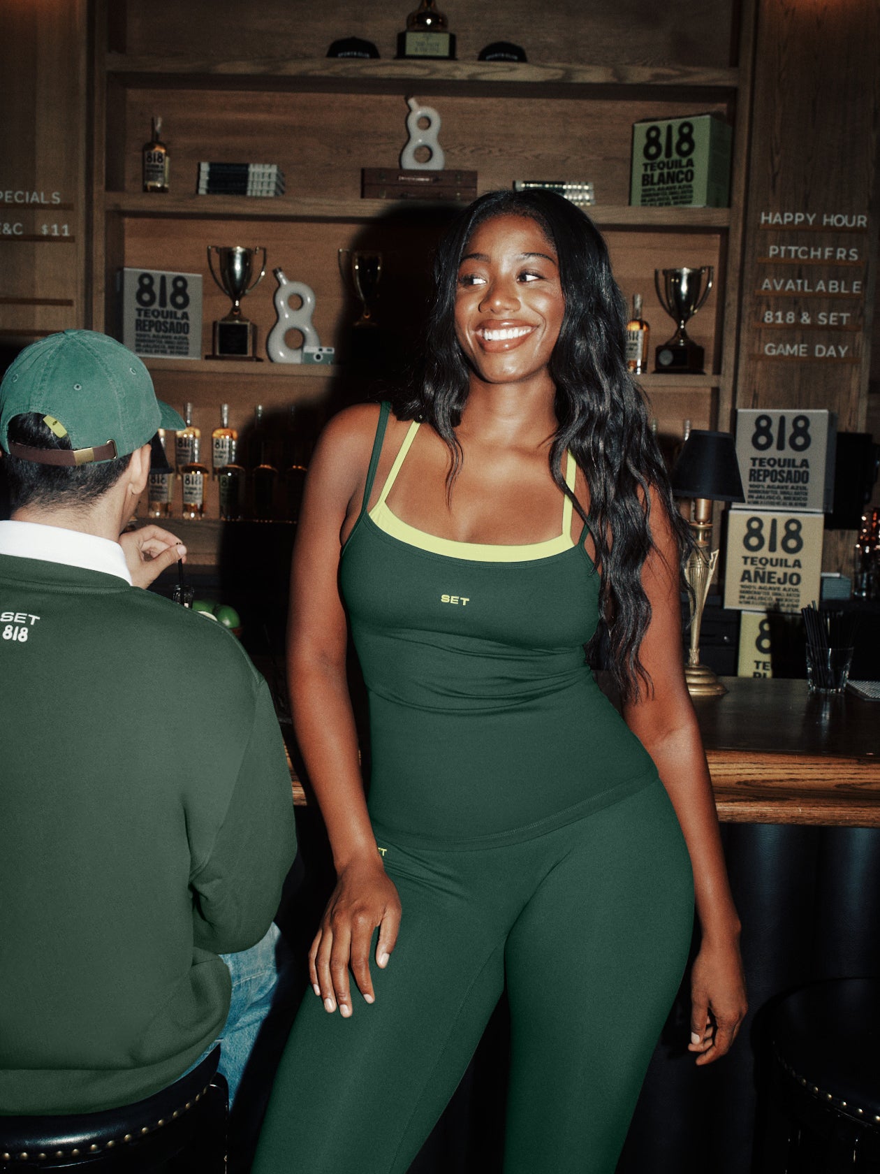 Woman in a green activewear outfit sitting in a bar with a man in a green cap.