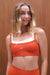 Woman wearing an orange sports bra with 'SET' branding against a wooden background <model-info>Grace wears a size XS and is 5'8", 25" waist, 32" bust.</model-info>