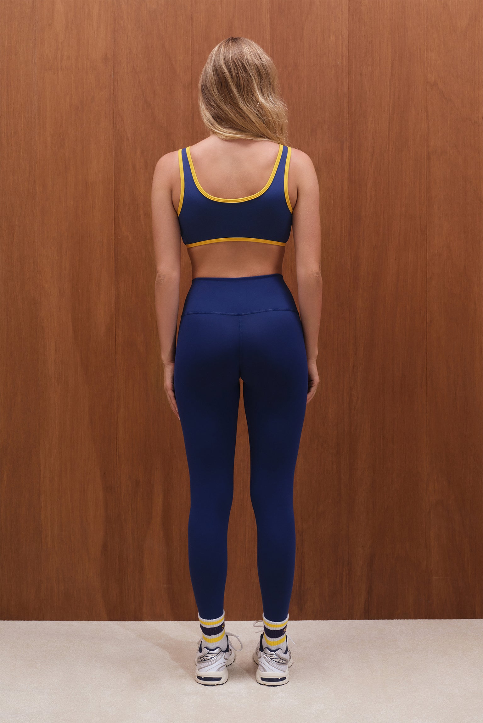 Person wearing a blue sports bra and leggings set with yellow accents against a wooden wall.