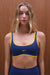 Woman wearing a blue sports bra with yellow trim against a wooden background <model-info>Grace wears a size XS and is 5'8", 25" waist, 32" bust.</model-info>