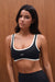 Woman wearing a black sports bra with white accents against a wooden background <model-info>Sydnee wears a size XS and is 5'8", 22” waist, 32BB bust.</model-info>