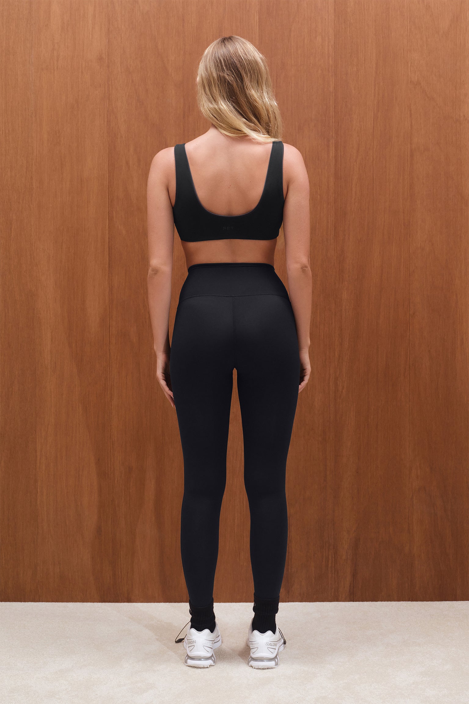 Person wearing a black sports bra and leggings set against a wooden wall.