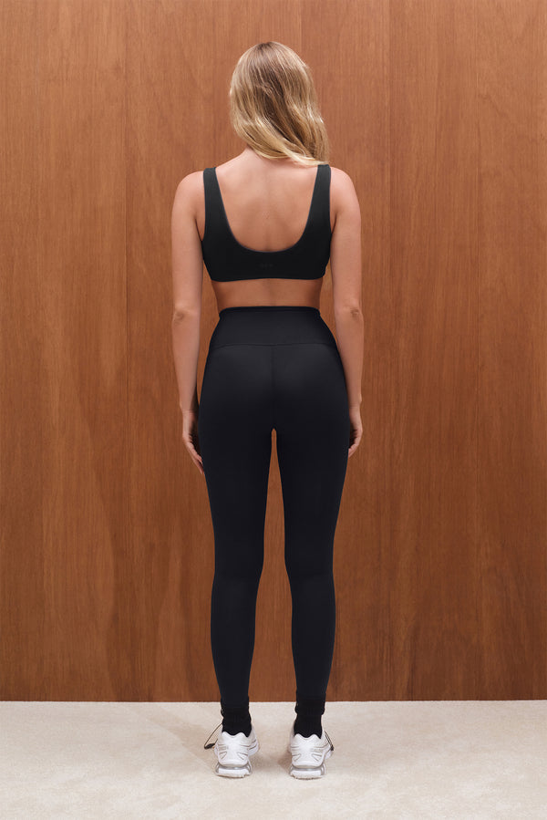 Person wearing a black sports bra and leggings set against a wooden wall.