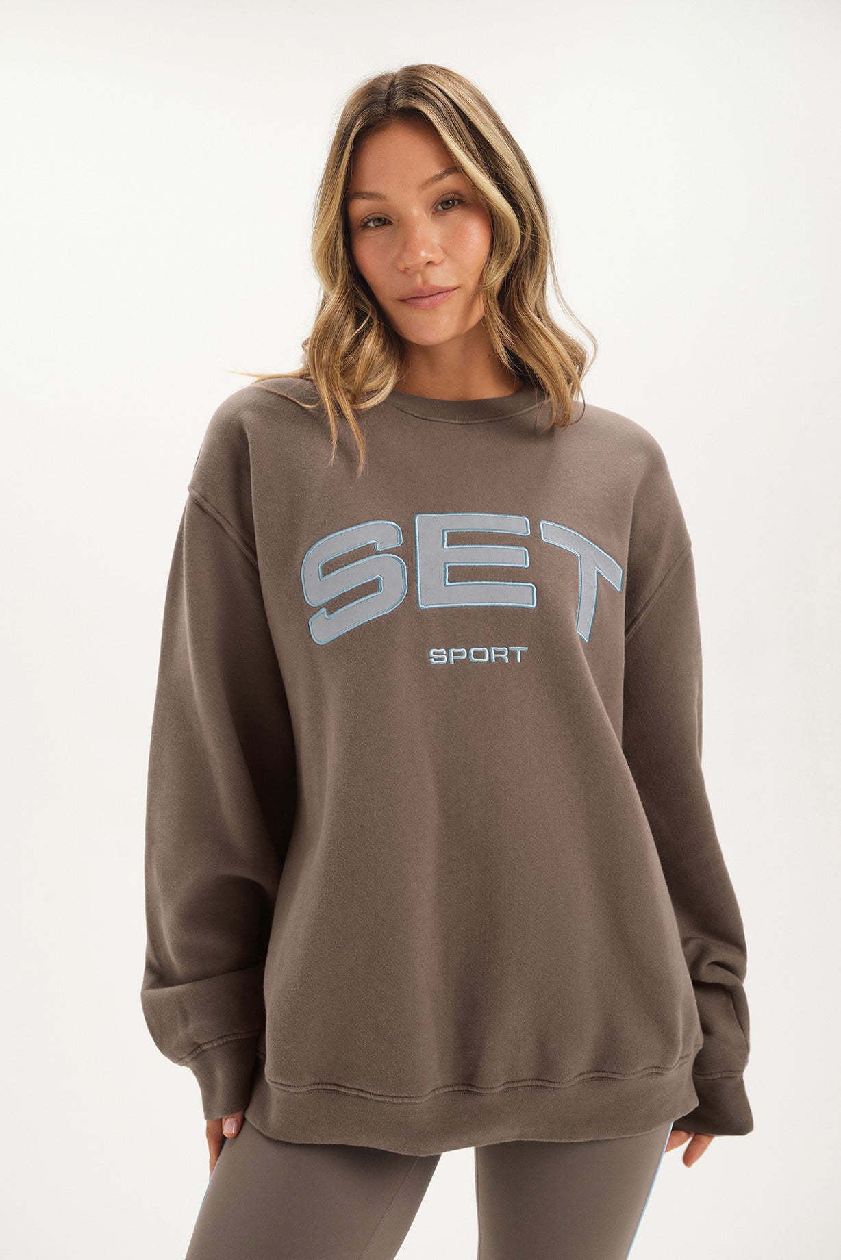 Woman wearing a brown sweatshirt with 'SET Sport' logo on a white background <model-info>Bre wears a size L and is 5’7”, 25½" waist, 33” bust.</model-info>