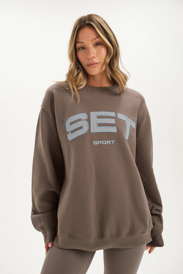Woman wearing a brown sweatshirt with 'SET Sport' logo on a white background 
