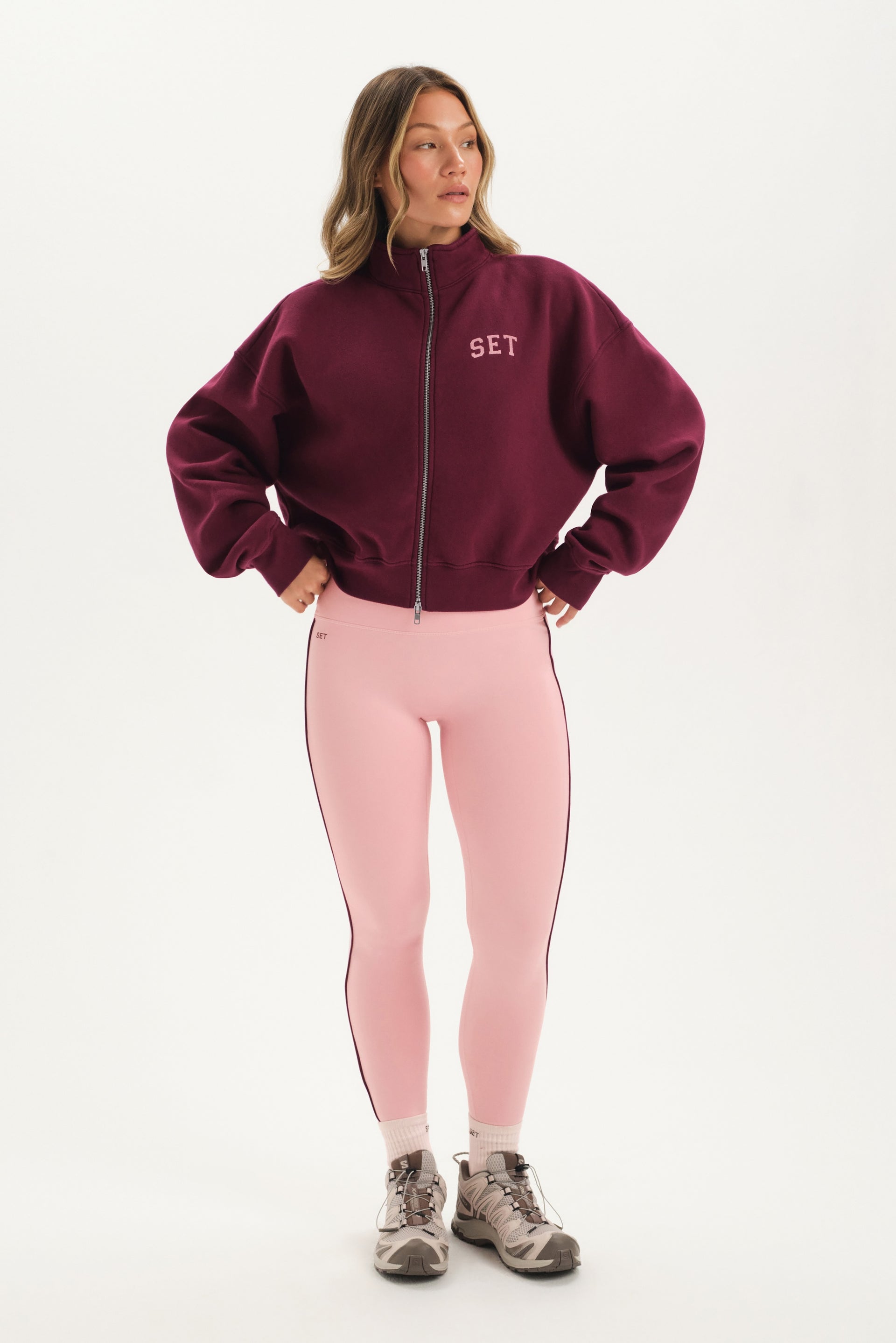 Woman wearing a burgundy jacket with 'SET' logo and pink leggings on a white background 
