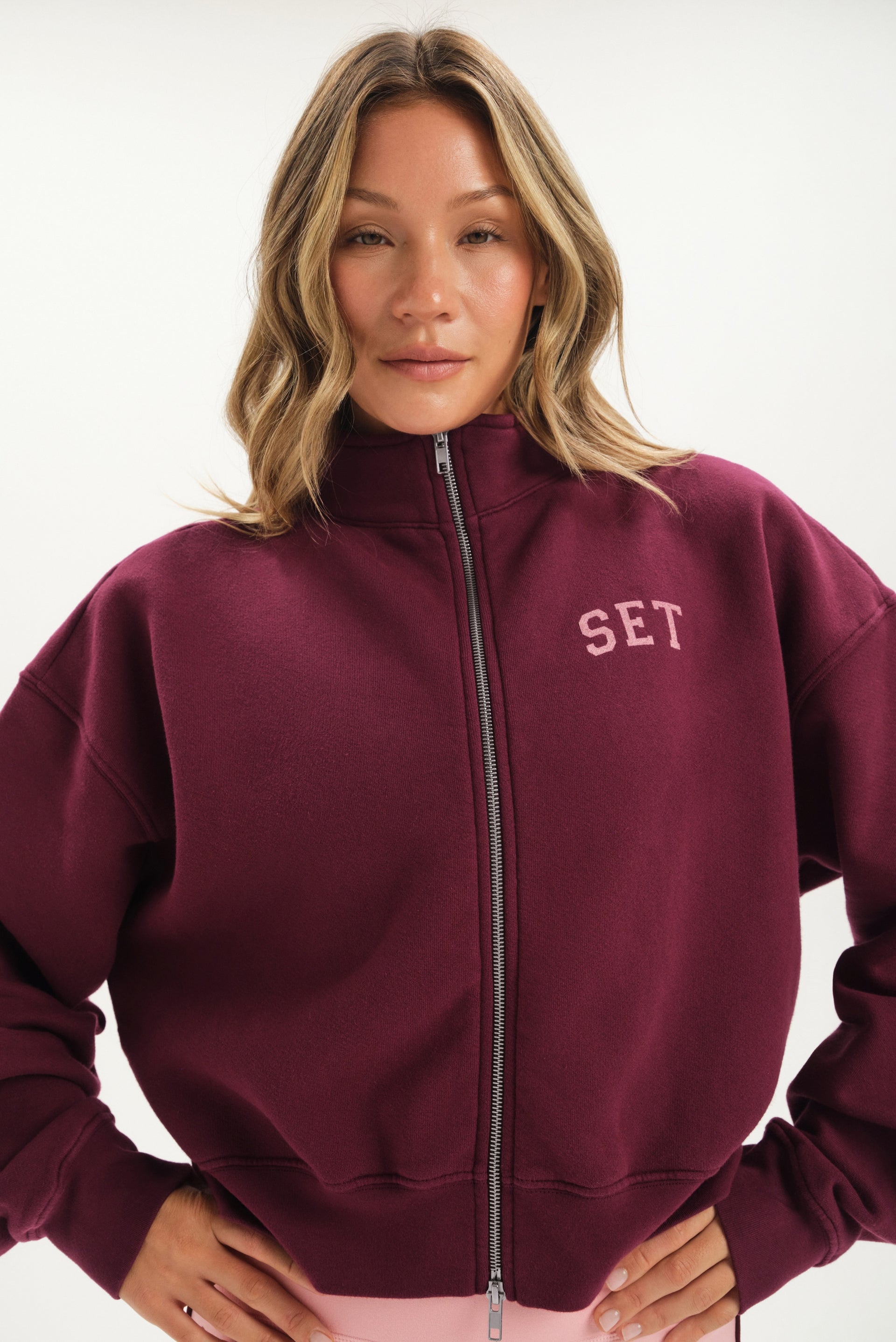 Woman wearing a maroon zip-up hoodie with 'SET' logo on a white background <model-info>Bre wears a size M and is 5’7”, 25½" waist, 33” bust.</model-info>
