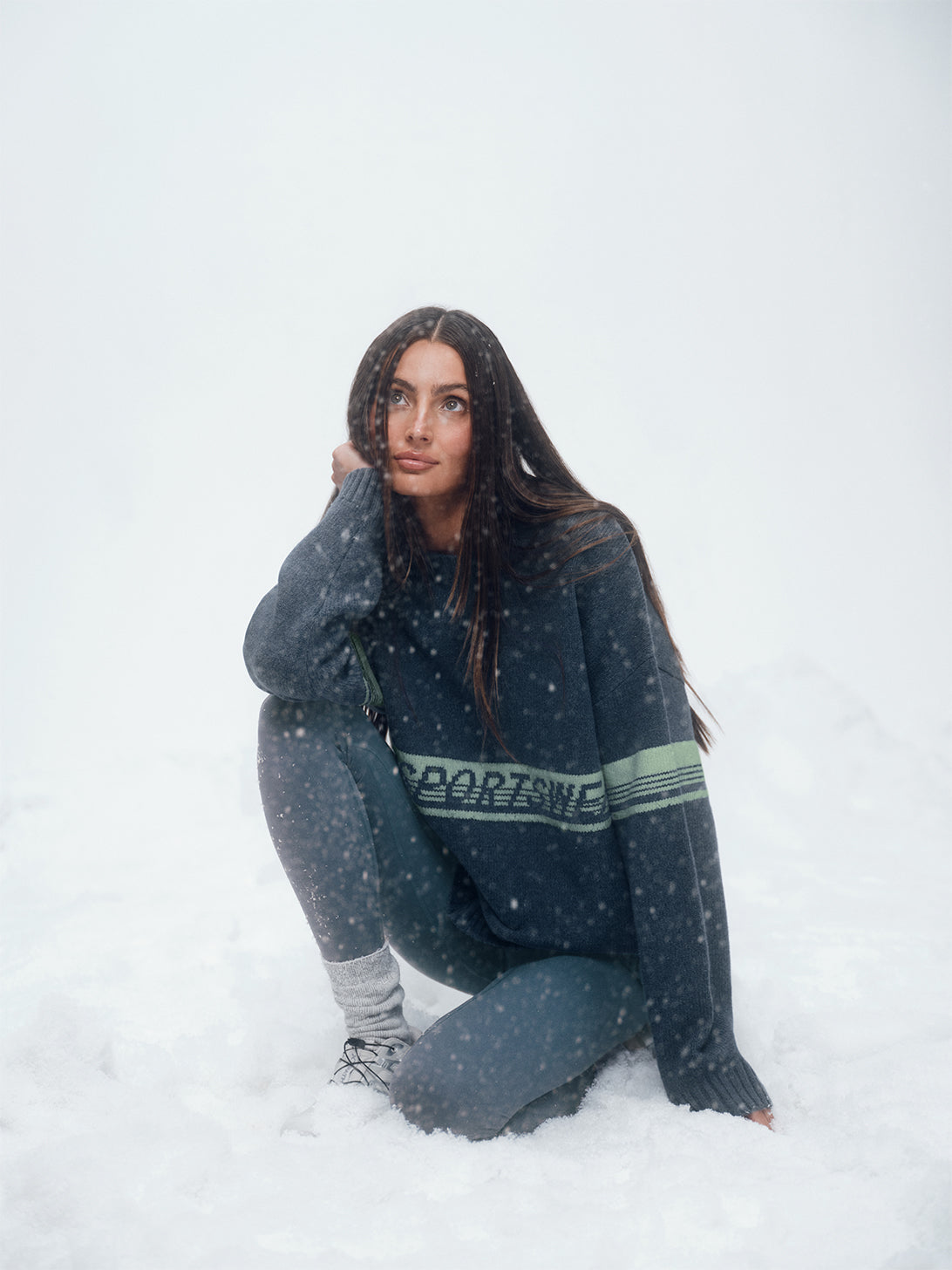 Person sitting in the snow wearing a dark sweater with text and leggings.