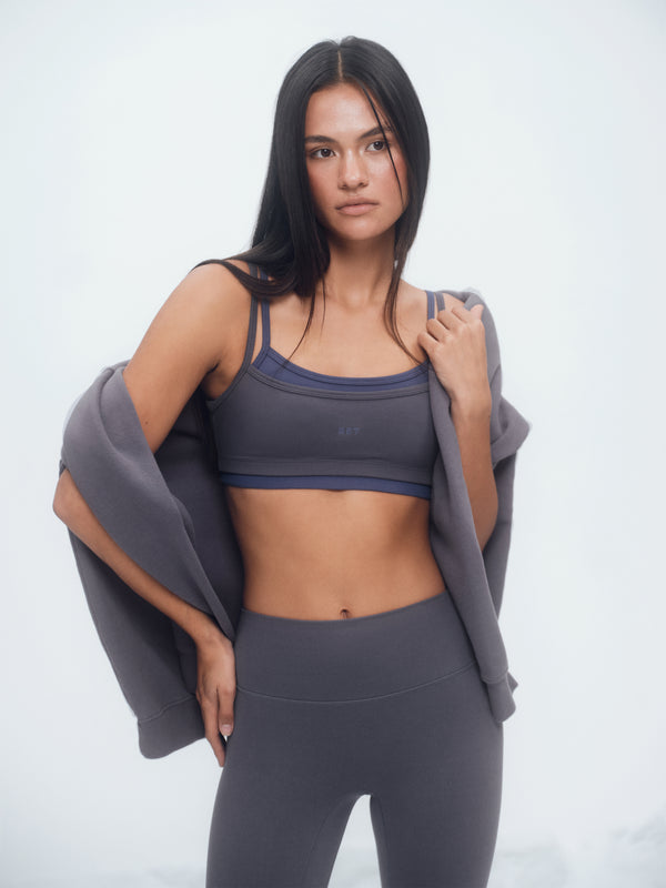 Woman wearing a gray sports bra and leggings with a gray jacket over her shoulder on a white background