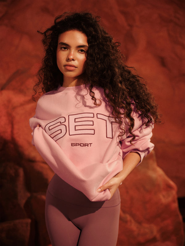 Person wearing a pink 'SET Sport' sweatshirt against a textured red-brown background