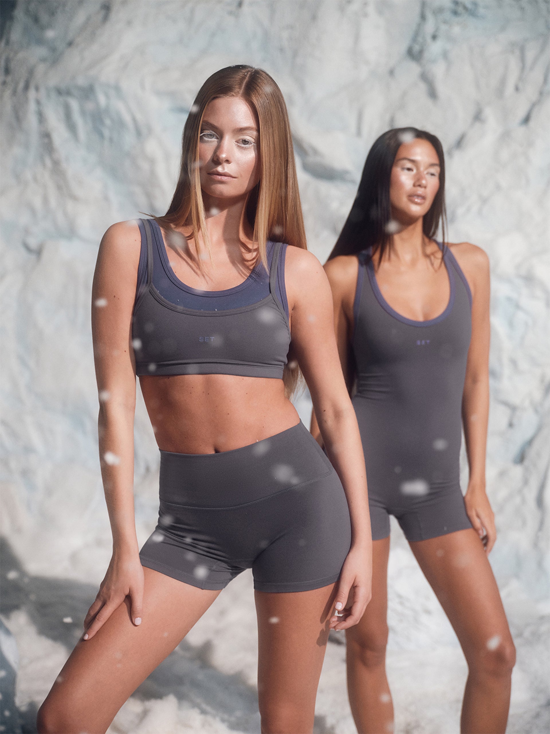 Two women wearing matching gray athletic outfits against a textured wall.