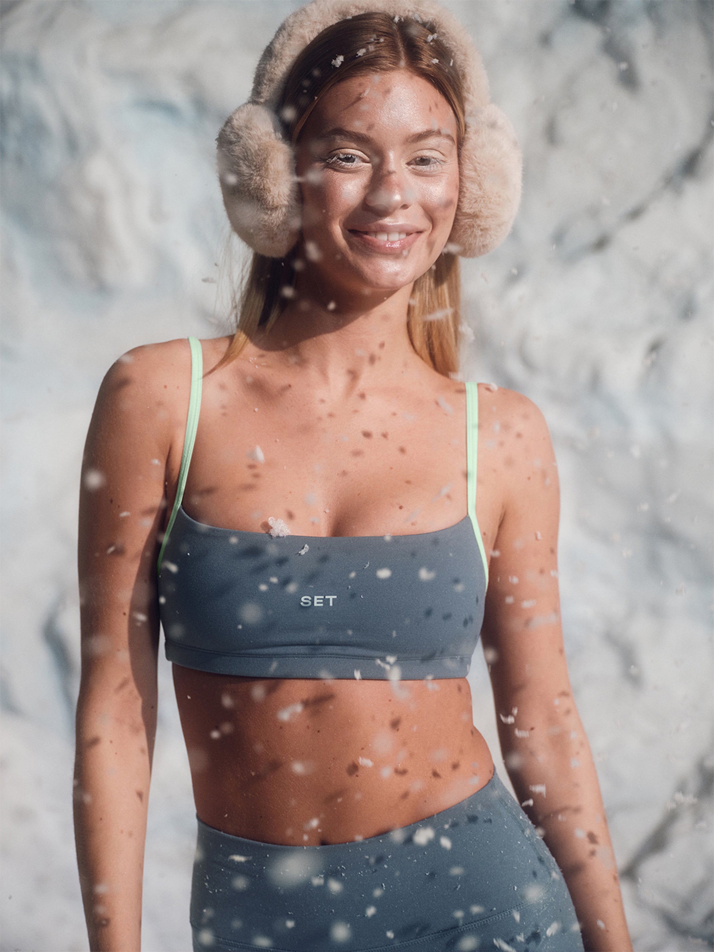 Woman wearing a blue sports bra and leggings with ear muffs in a snowy setting.