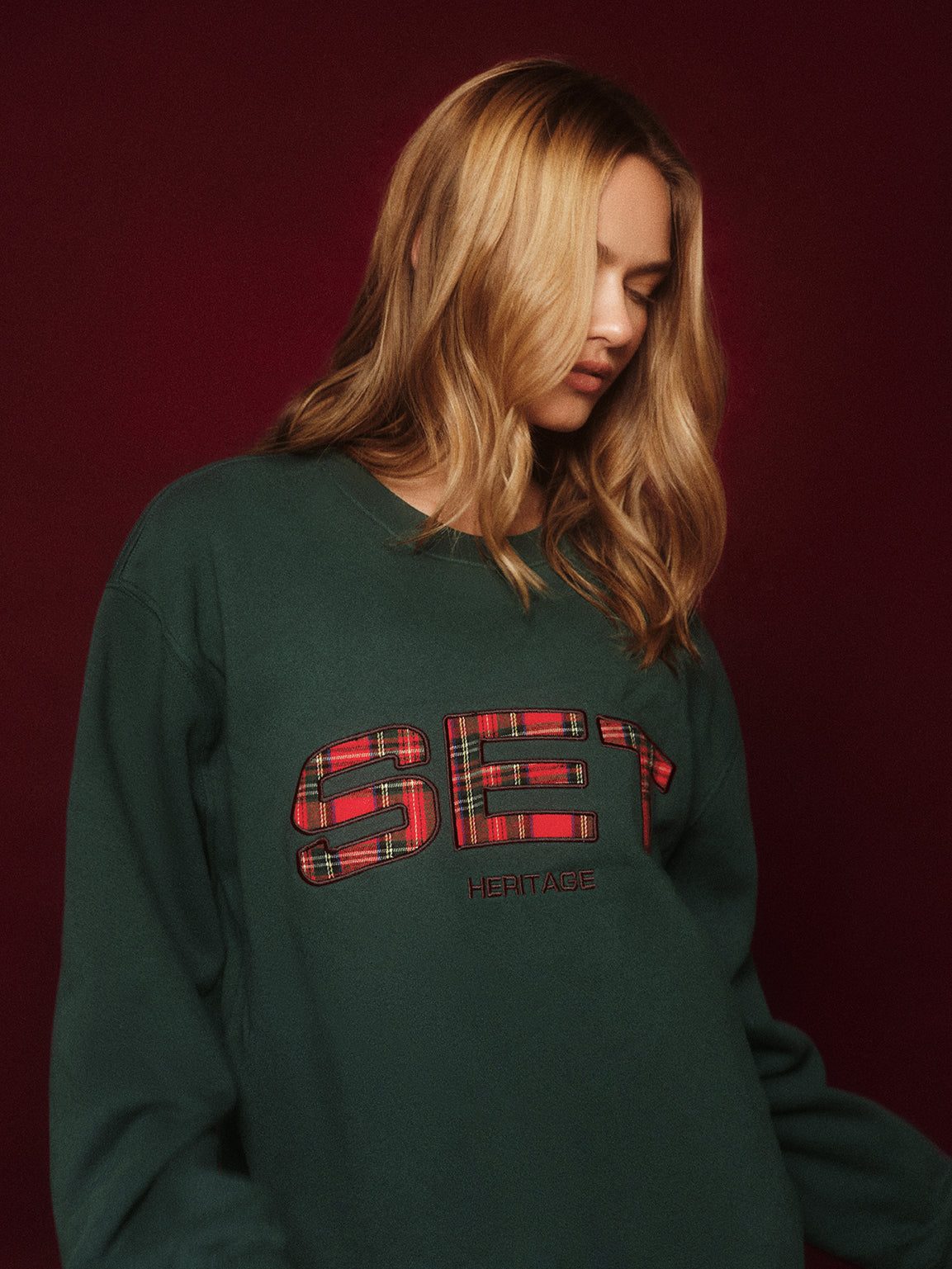 Person wearing a green sweatshirt with red plaid lettering against a dark red background 