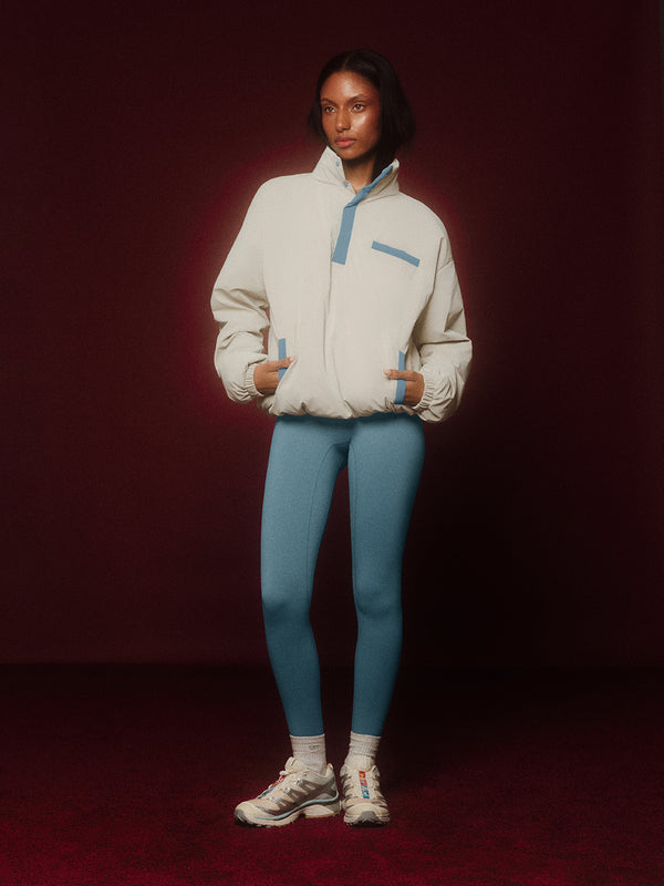 Person wearing a beige jacket with blue accents and blue leggings on a dark red background