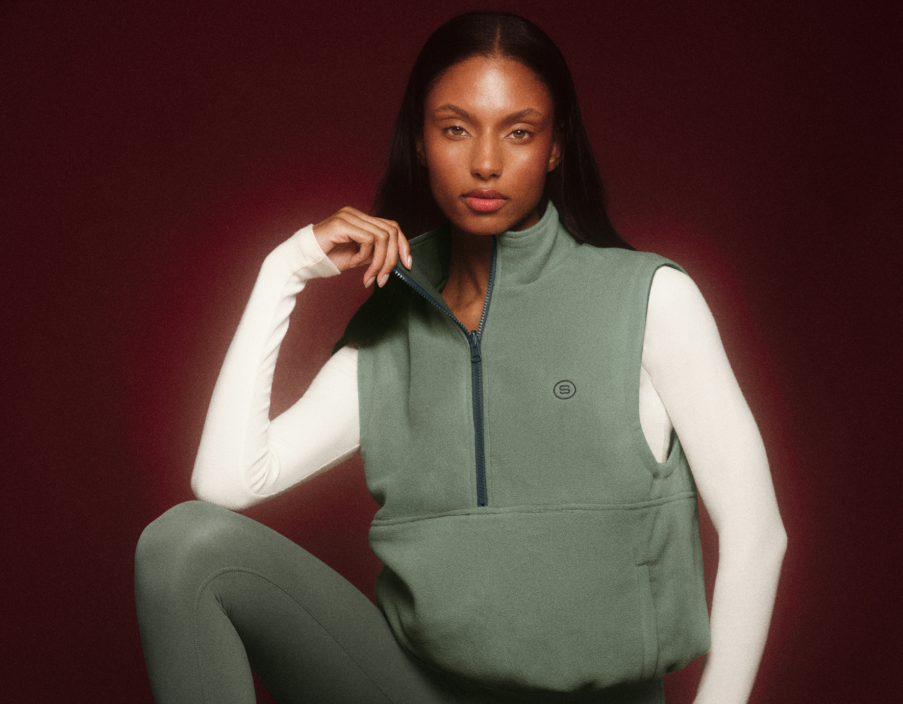 Woman wearing a green vest over a white long-sleeve shirt against a red background