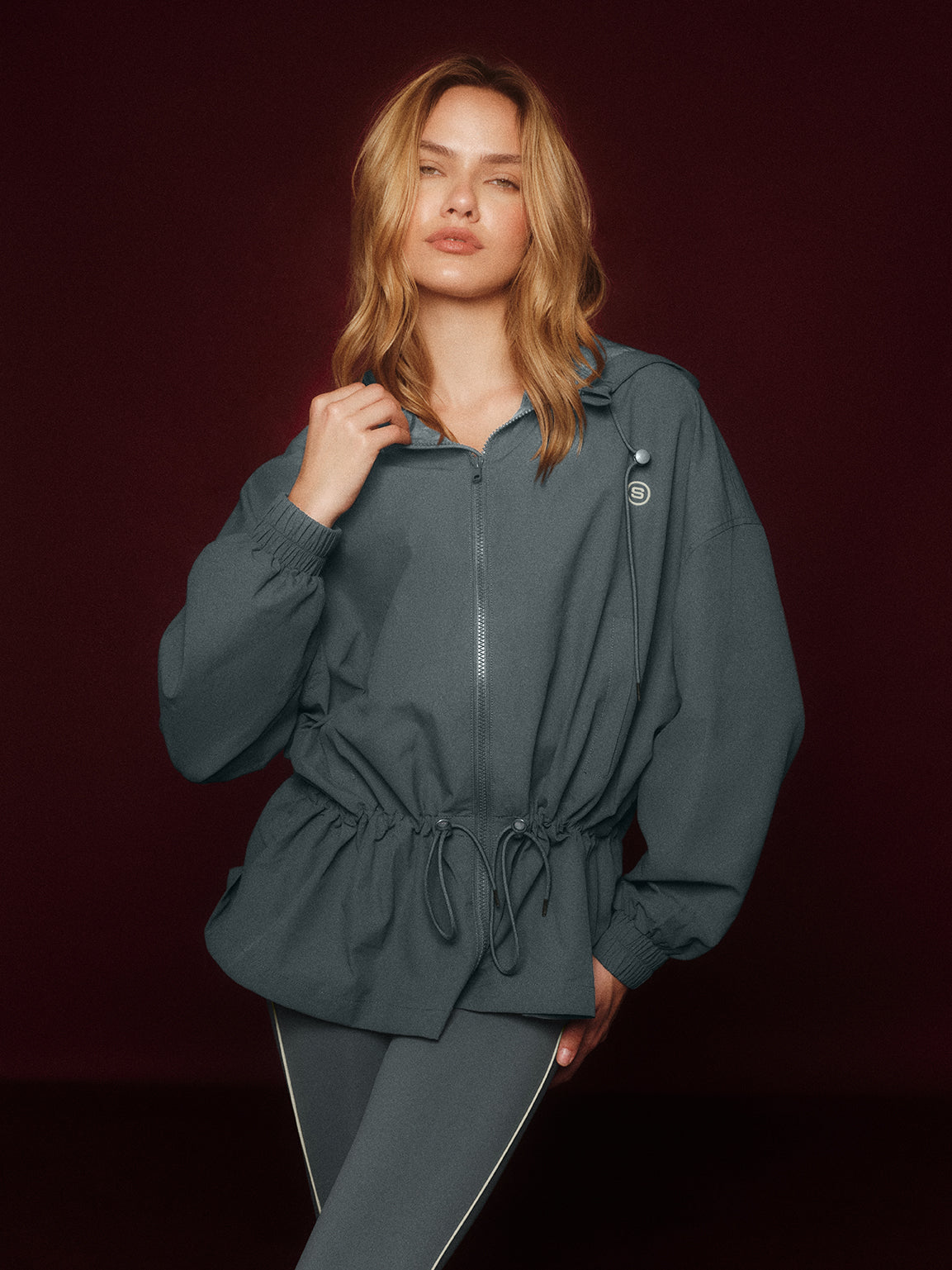 Woman wearing a dark gray cinched windbreaker against a dark background