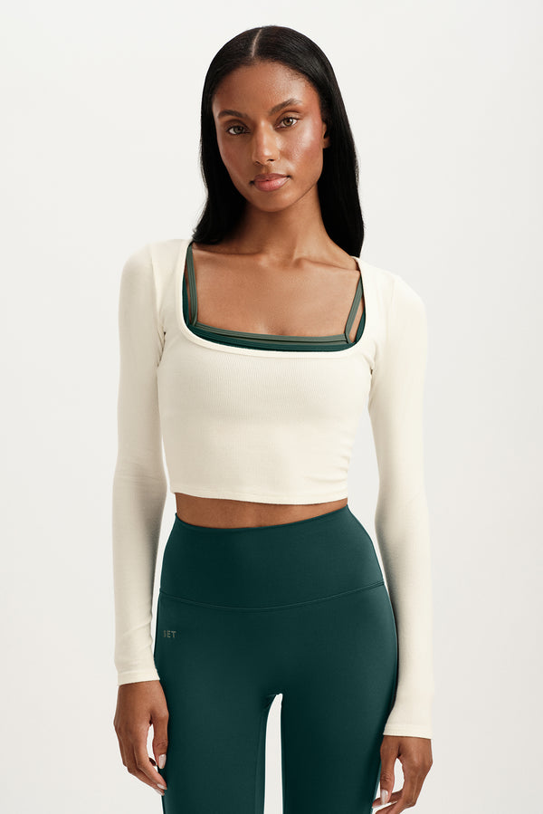 Woman wearing a white long-sleeve crop top and dark green leggings on a light background 