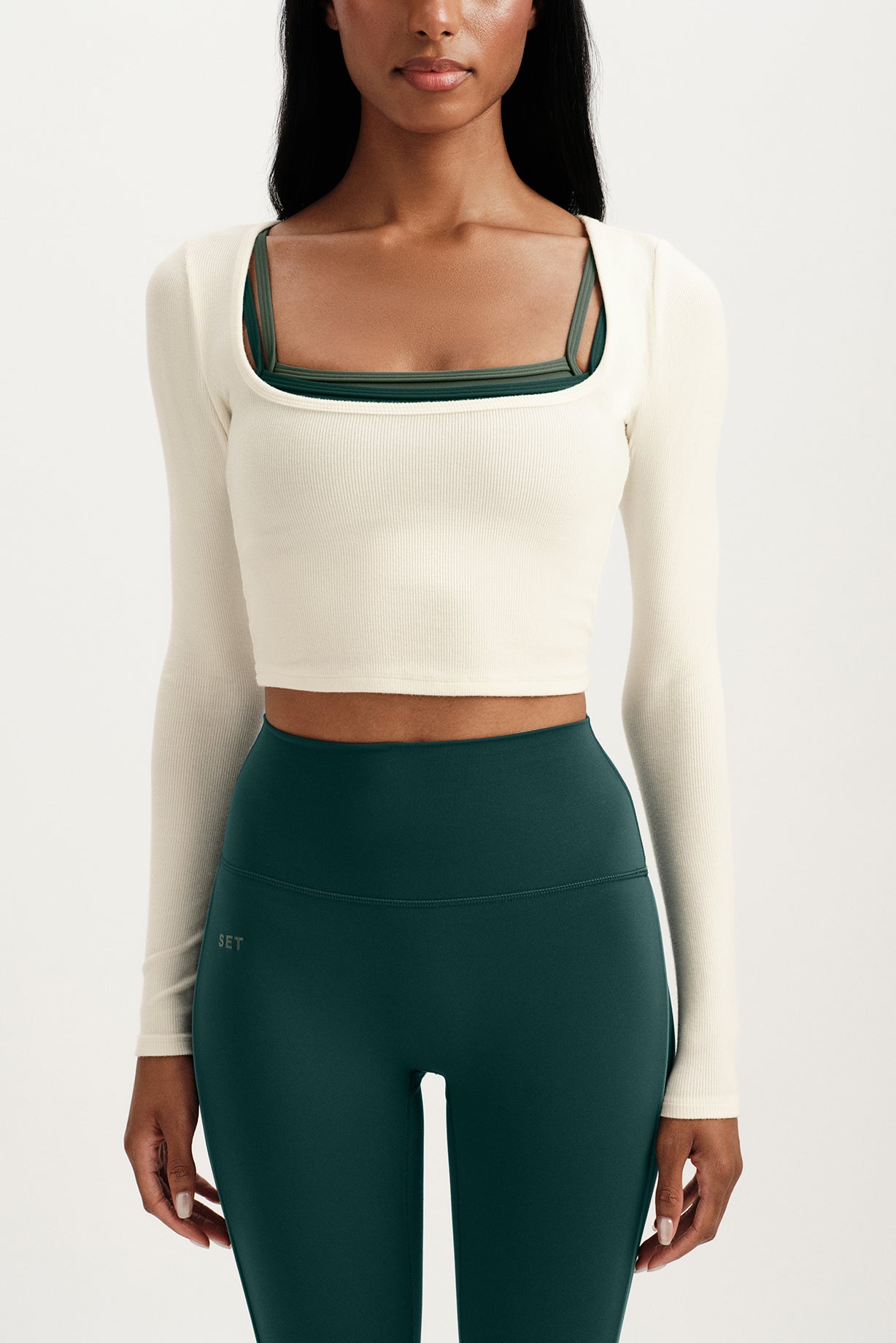 Person wearing a white long-sleeve crop top with green neckline and dark green leggings on a white background