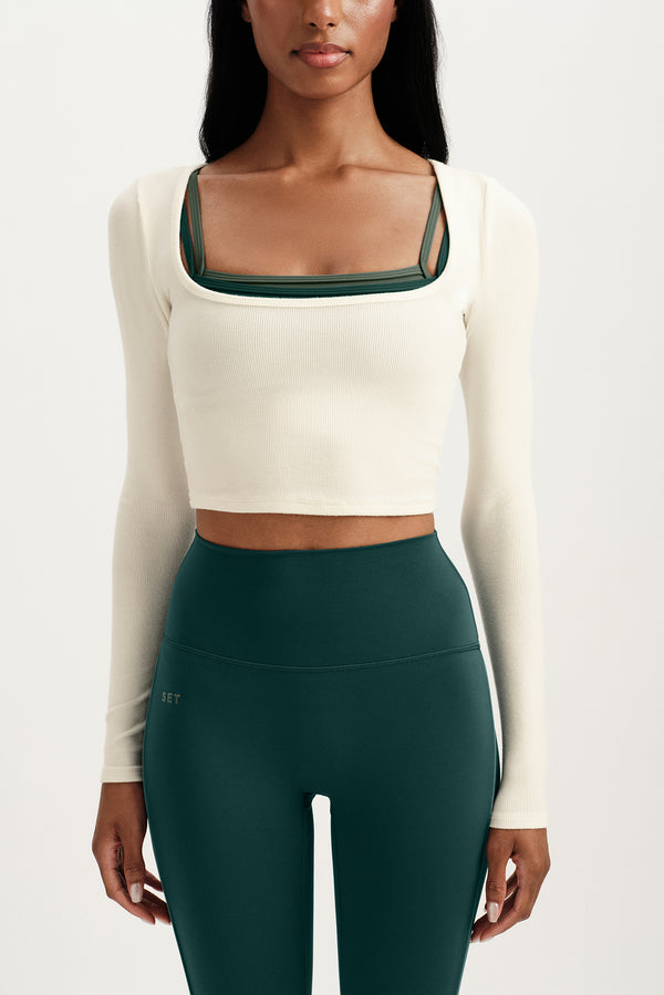 Person wearing a white long-sleeve crop top with green neckline and dark green leggings on a white background