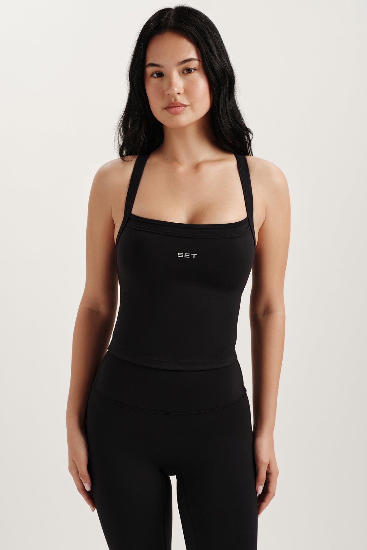 Woman wearing a black tank top with 'SET' logo on a light background <model-info>Jasmine wears a size S and is 5’8, 25” waist, 32C bust.</model-info> 