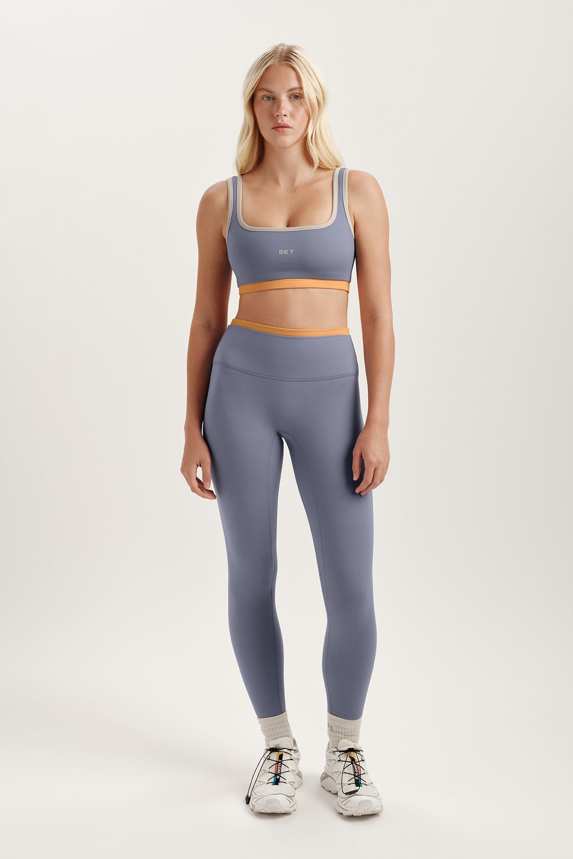 Woman wearing a purple sports bra and leggings with orange accents on a white background