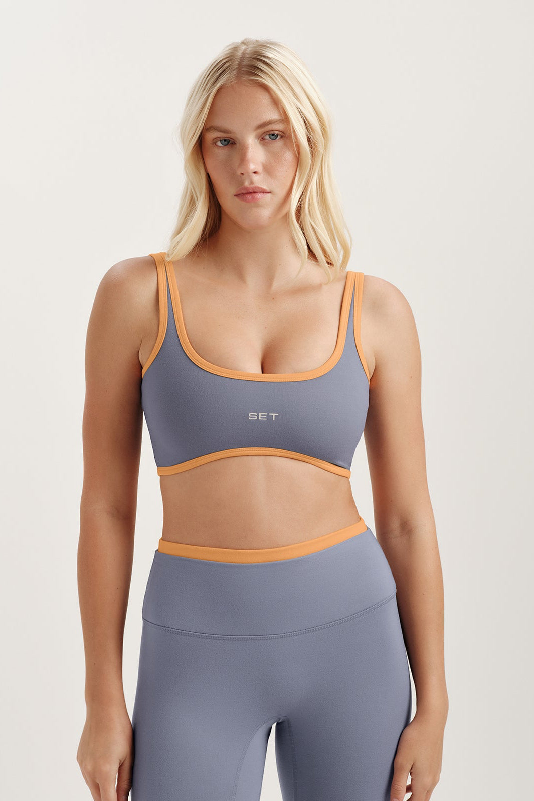 Woman wearing a purple sports bra and leggings with orange accents on a white background <model-info>Kenzie wears a size S and is 5’6, 25” waist, 33” bust.</model-info> 