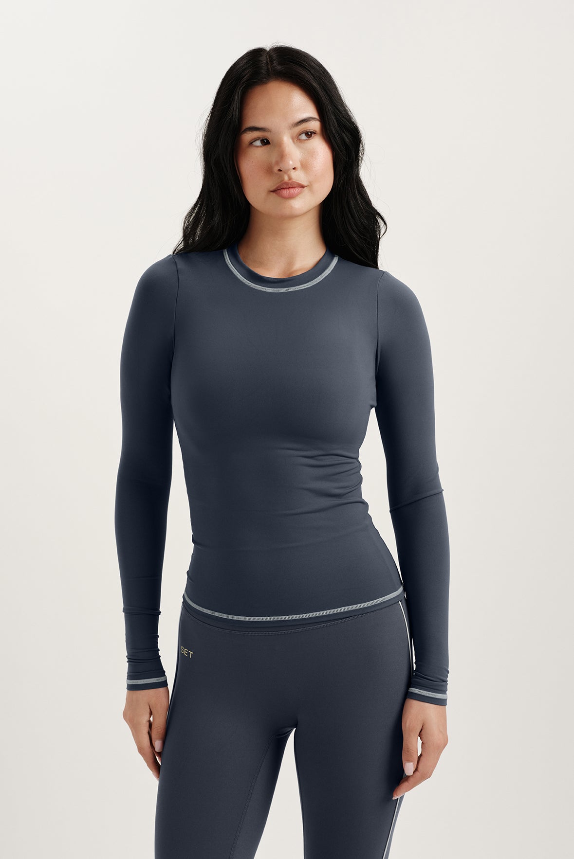 Woman wearing a dark gray long-sleeve top and leggings on a light background <model-info>Jasmine wears a size S and is 5’8, 25” waist, 32C bust.</model-info> 