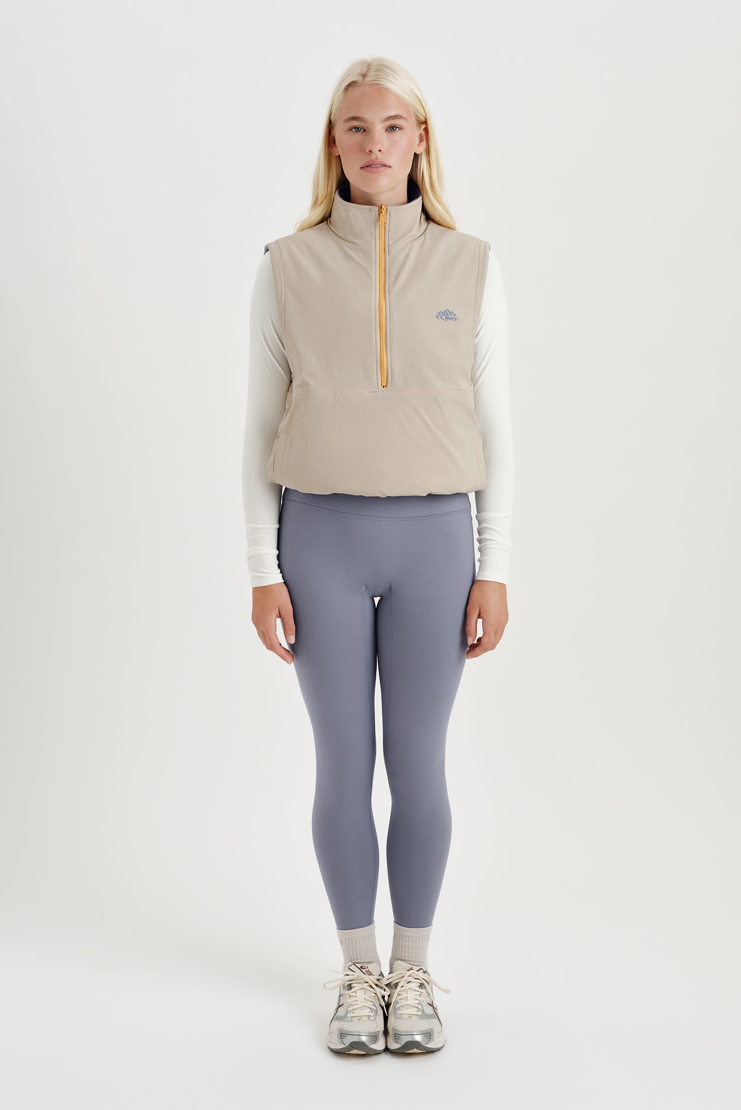 Woman wearing a beige insulated vest and gray leggings on a white background