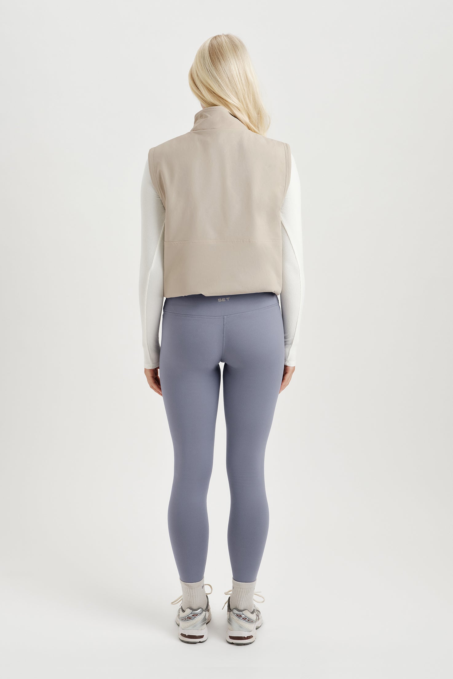 Person wearing a beige vest, white shirt, gray leggings, and athletic shoes on a white background