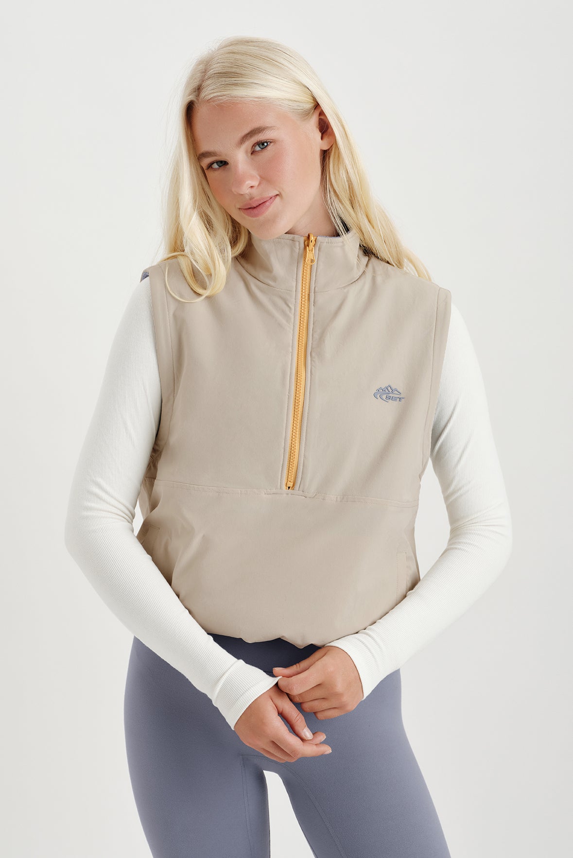 Woman wearing a beige vest with a brand logo on a light gray background 