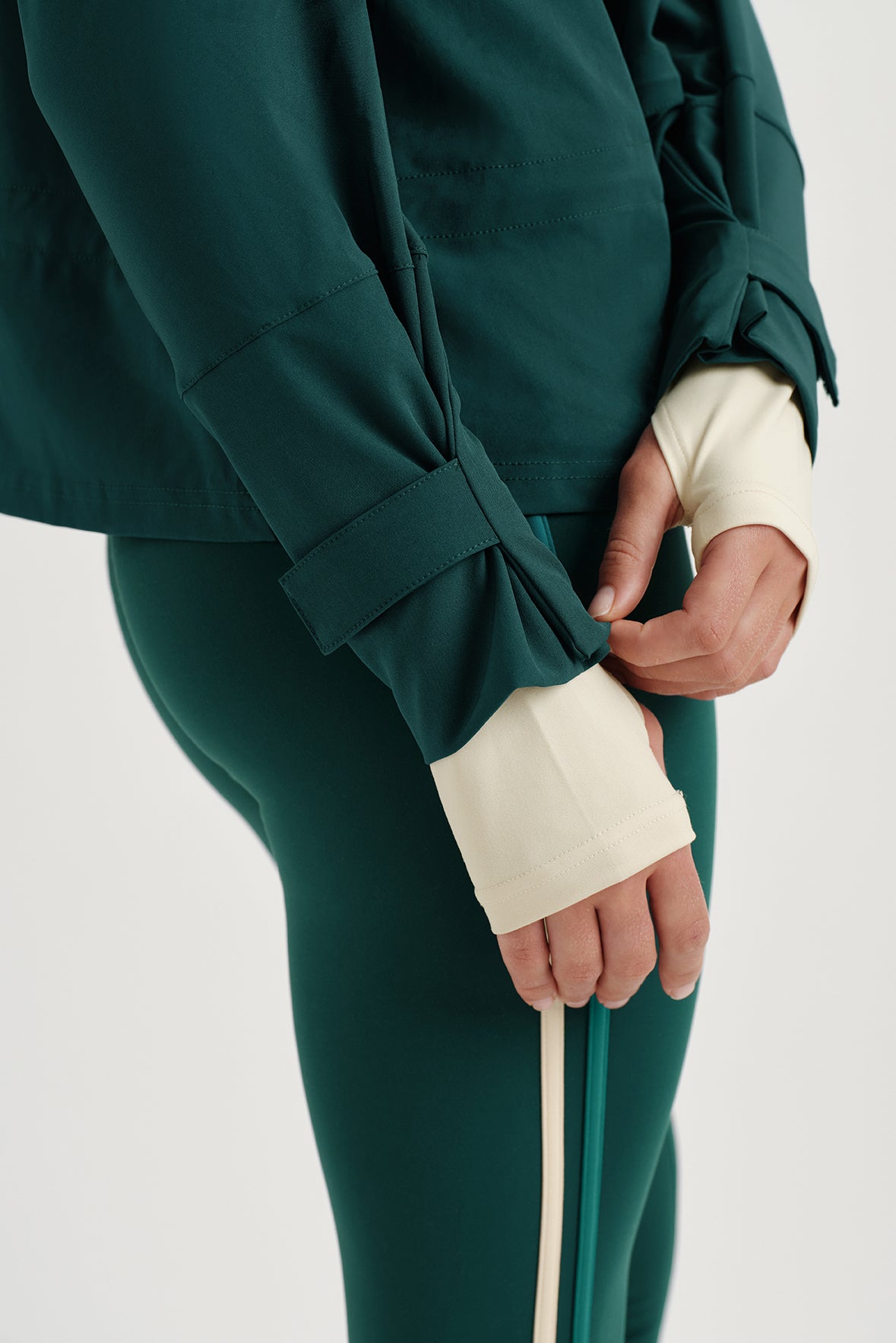 Person wearing a green outfit with a white shirt underneath with thumbholes on a light gray background