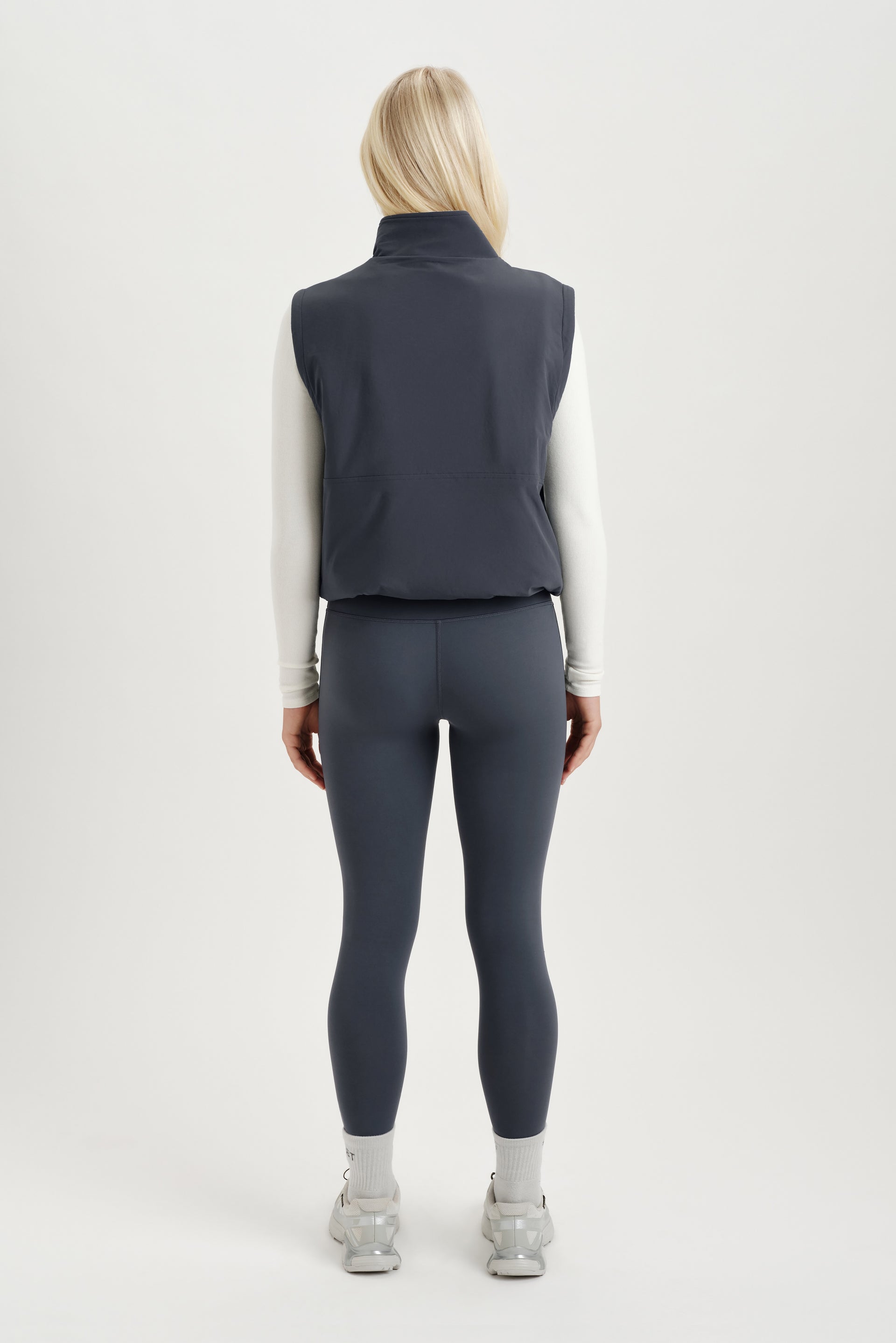 Woman turned around wearing a white long sleeve and gray vest on a white background  